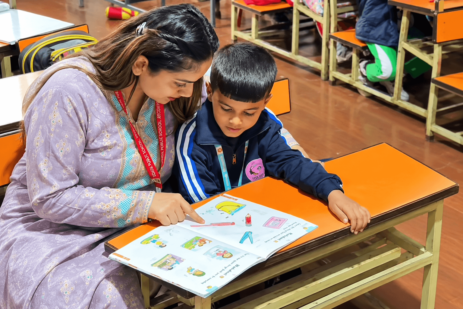 A teacher sitting beside a young student, helping him read from a colourful book at a classroom desk